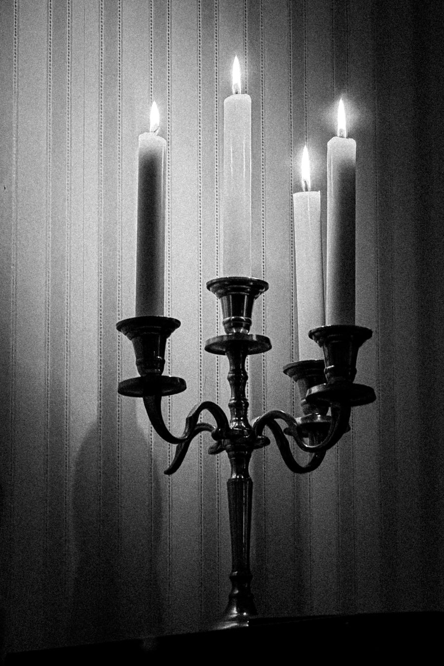 A black and white image of an ornate metal candelabra holding four lit candles of varying heights, their flames glowing against a striped, softly lit wallpaper background. The candelabra’s curved arms and dark metal contrast with the pale wax of the candles.