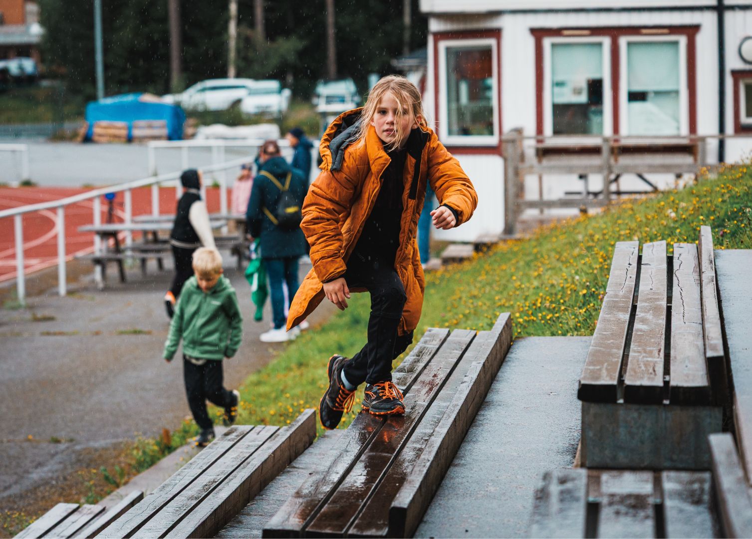 A child in an orange puffer coat balances while walking along a wet wooden bench at an outdoor athletics stadium, arms slightly raised. Another child in a green jacket runs nearby, with adults and a running track visible in the background under rainy conditions. A white and red building stands behind a grassy slope dotted with yellow wildflowers.