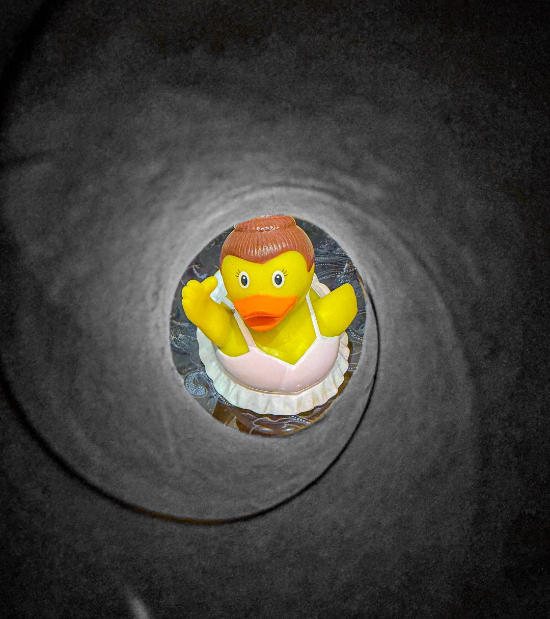 A rubber duck figure dressed in a white apron and orange headband peers up from inside a dark, tunnel-like drain opening, photographed with an ultra-wide lens that creates a strong vignette effect.