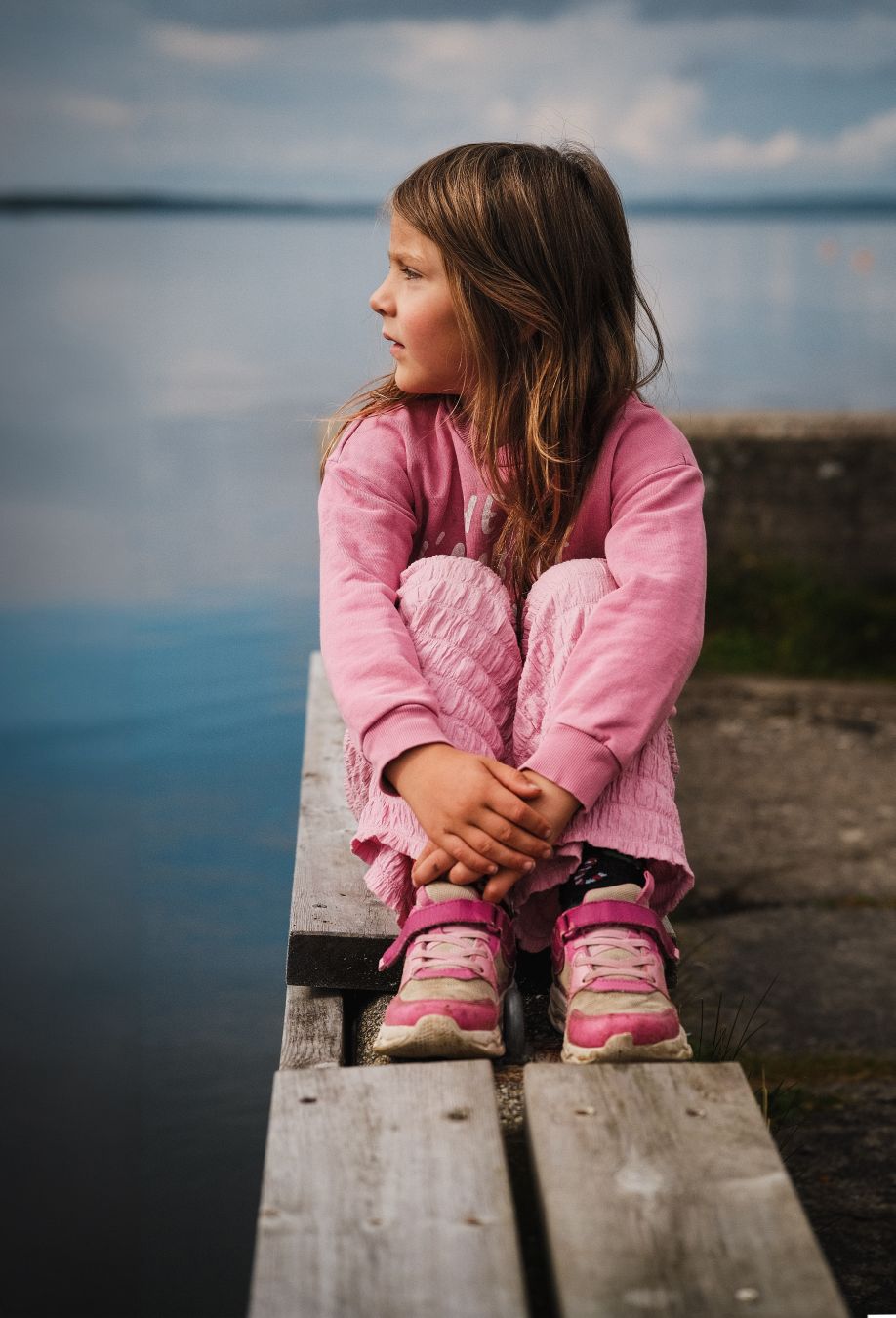 A young girl in a pink outfit sits on a wooden dock with her arms wrapped around her knees, gazing thoughtfully out over a calm lake. Soft blue tones from the water and sky dominate the background, with a rocky shoreline visible on the right.