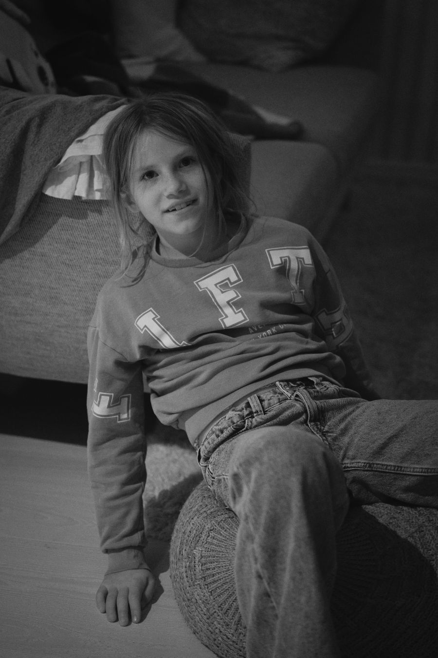 A black and white portrait of a child sitting sideways on a couch, one leg propped up, smiling slightly toward the camera. Soft indoor lighting creates a relaxed, candid atmosphere in the dim living room setting.