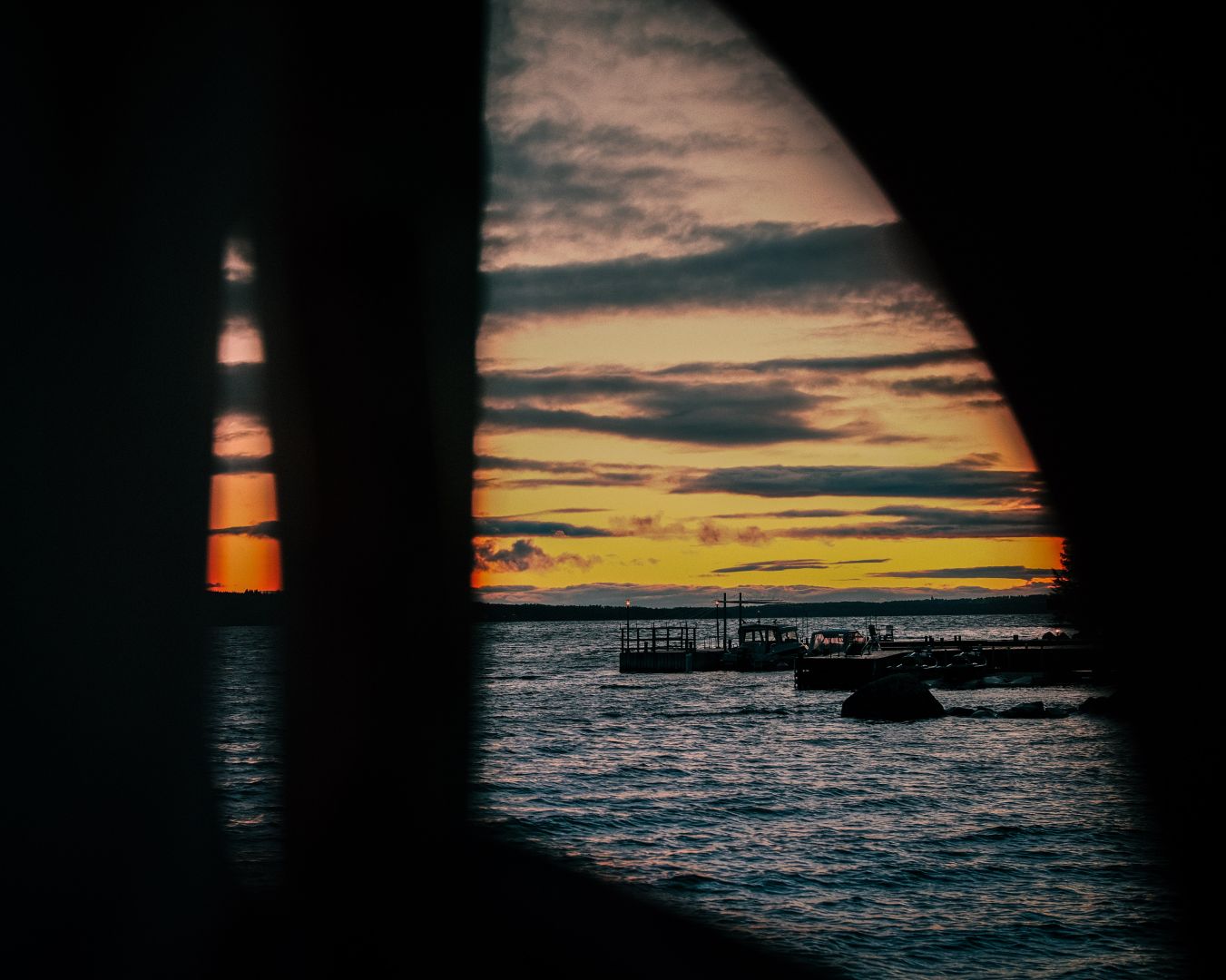 A sunset over a lake is framed through a dark, curved gap between structures, with warm orange and yellow light filling the cloudy sky above the water. A small dock with boats is visible in the distance, silhouetted against the glowing horizon.