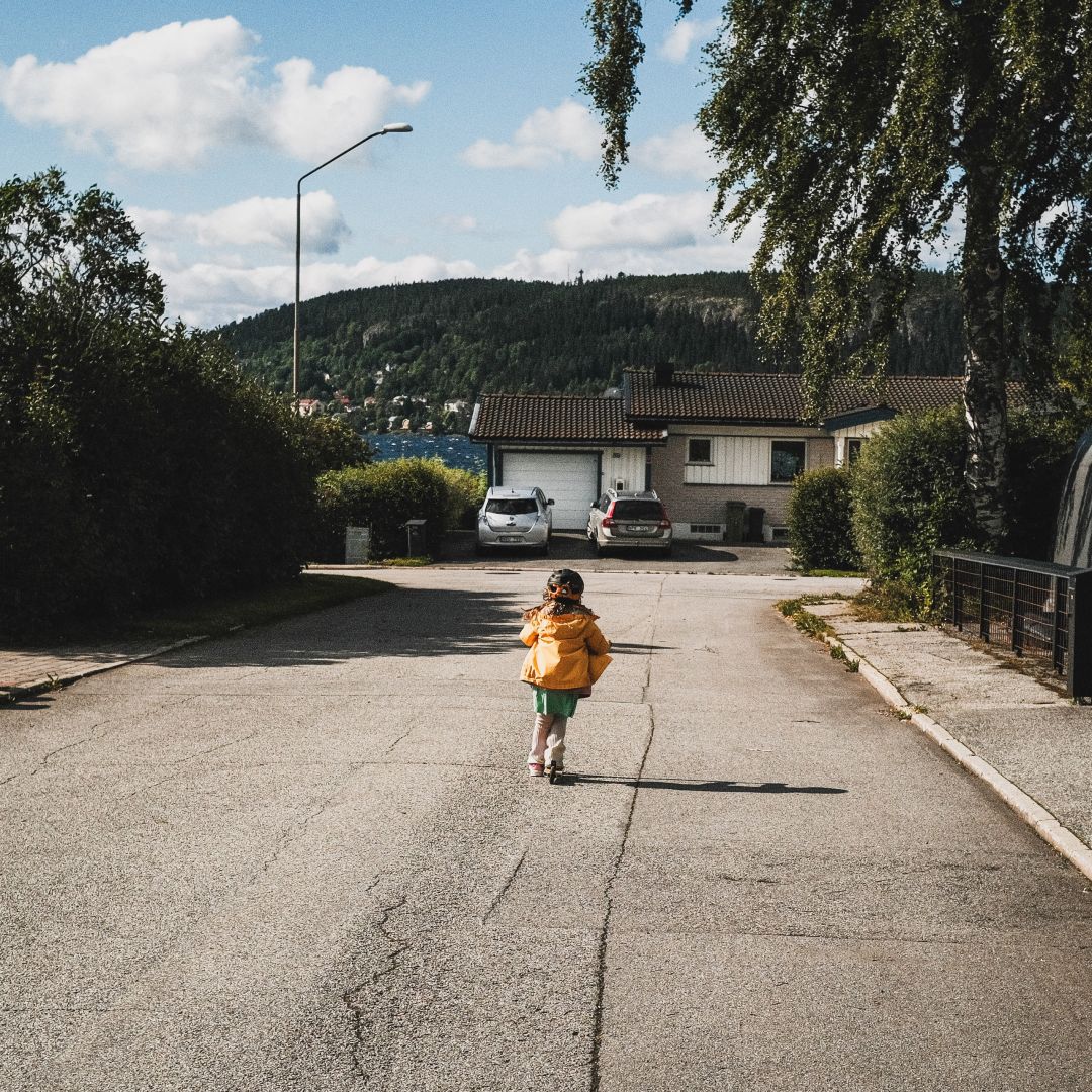 A small child wearing a helmet and orange jacket rides a scooter away from the camera down a residential asphalt street, past parked cars and houses. Tall birch trees frame the sides of the road, with a wooded hillside and a glimpse of lake water visible in the background under a partly cloudy sky.