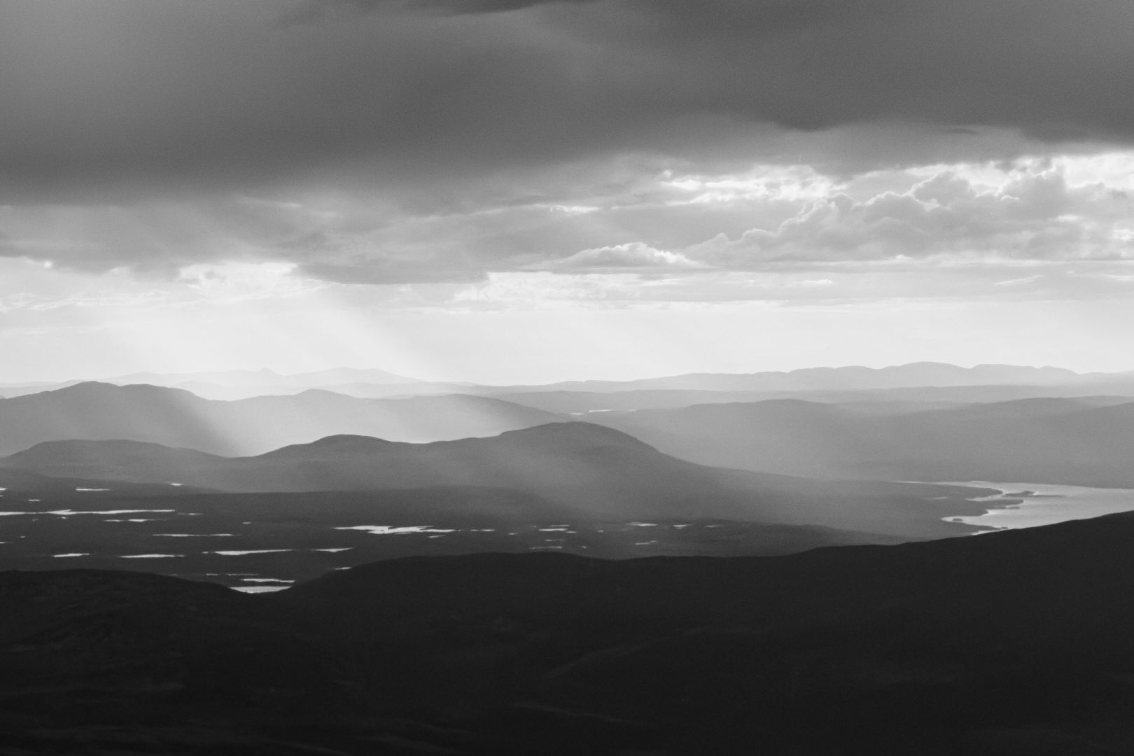 A black and white landscape image of layered mountain ridges fading into the distance under a dramatic sky, with sunbeams breaking through heavy clouds and illuminating patches of the terrain below, including a winding river or lake.