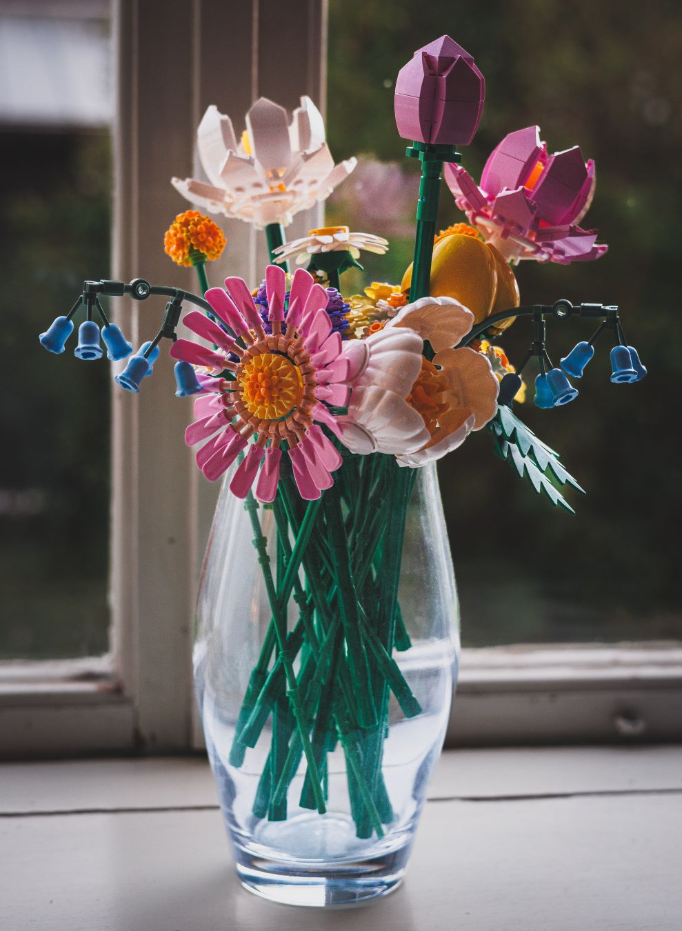A bouquet of LEGO flowers in bright pink, orange, purple, and blue arranges in a clear glass vase filled with water, standing on a windowsill. The intricate plastic blooms mimic real flowers like daisies, tulips, and bluebells, with soft daylight and a blurred outdoor view behind them.
