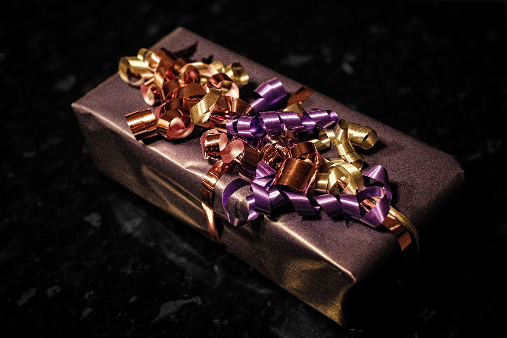 A close-up of a wrapped gift box in dark purple paper, topped with an elaborate bundle of curled ribbons in gold, bronze, and purple tones. The dark, textured background makes the metallic ribbons stand out sharply.