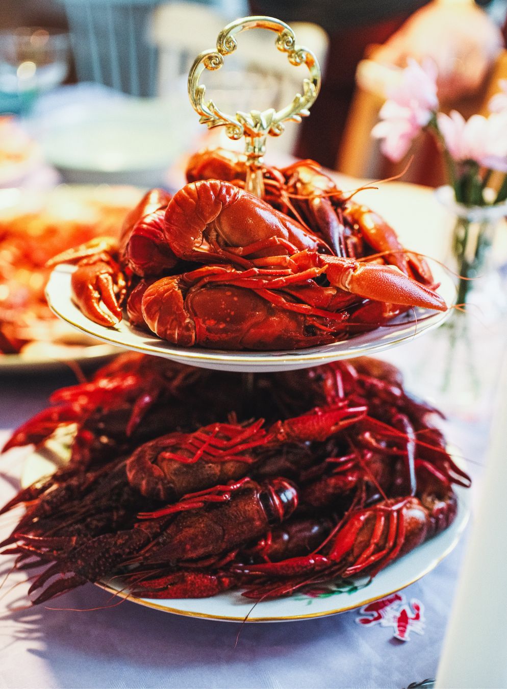 A two-tiered serving stand piled high with cooked crayfish, their bright red shells glistening under warm light. The stand is decorated with an ornate gold handle, and pink flowers are visible in a vase in the softly blurred background.