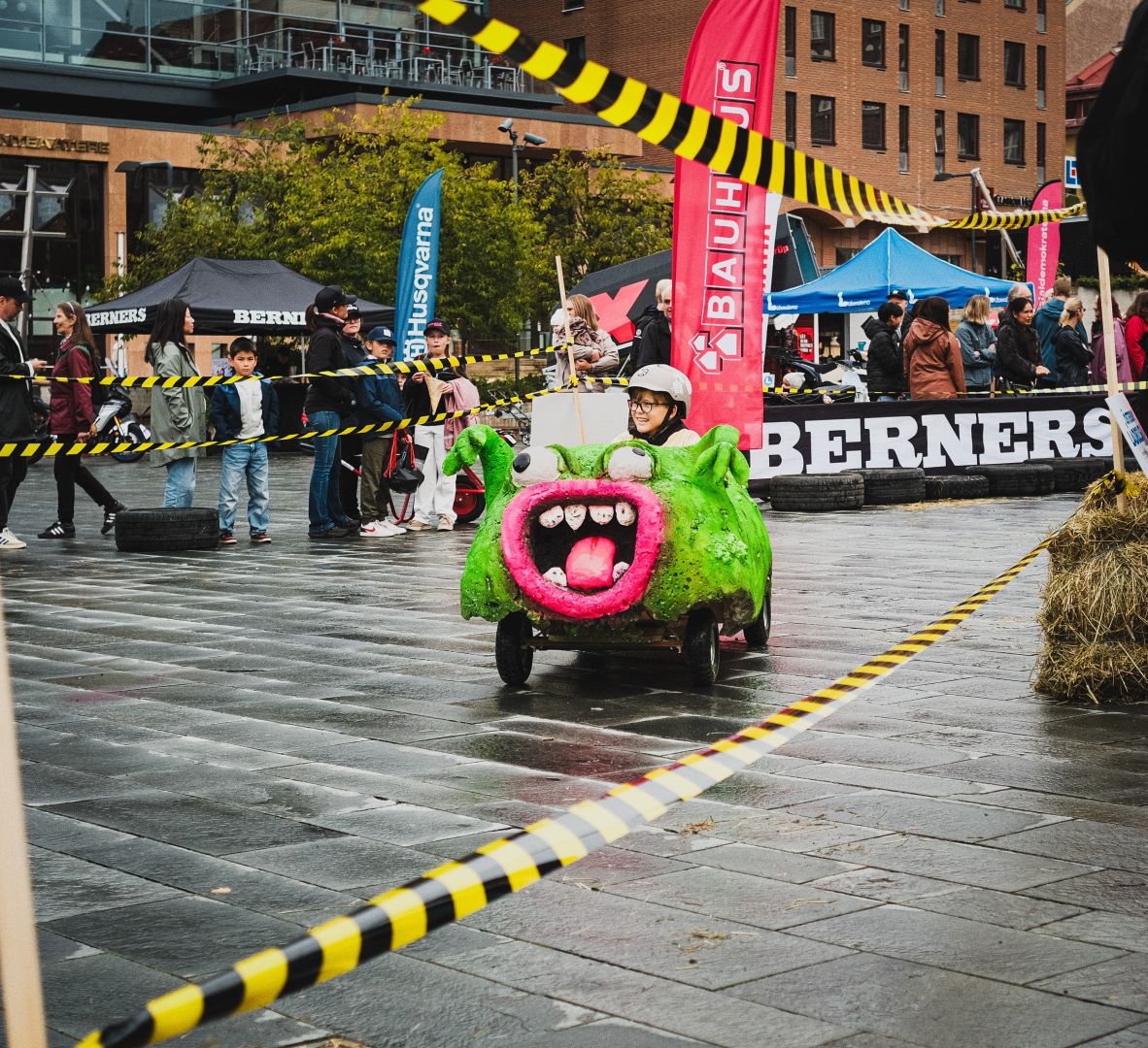 A child wearing a helmet drives a homemade soapbox cart shaped like a green monster with big eyes and a pink mouth, racing across a cobblestone city square lined with caution tape. Spectators, tents, and sponsor banners including Husqvarna and Bauhaus fill the background near a brick building.