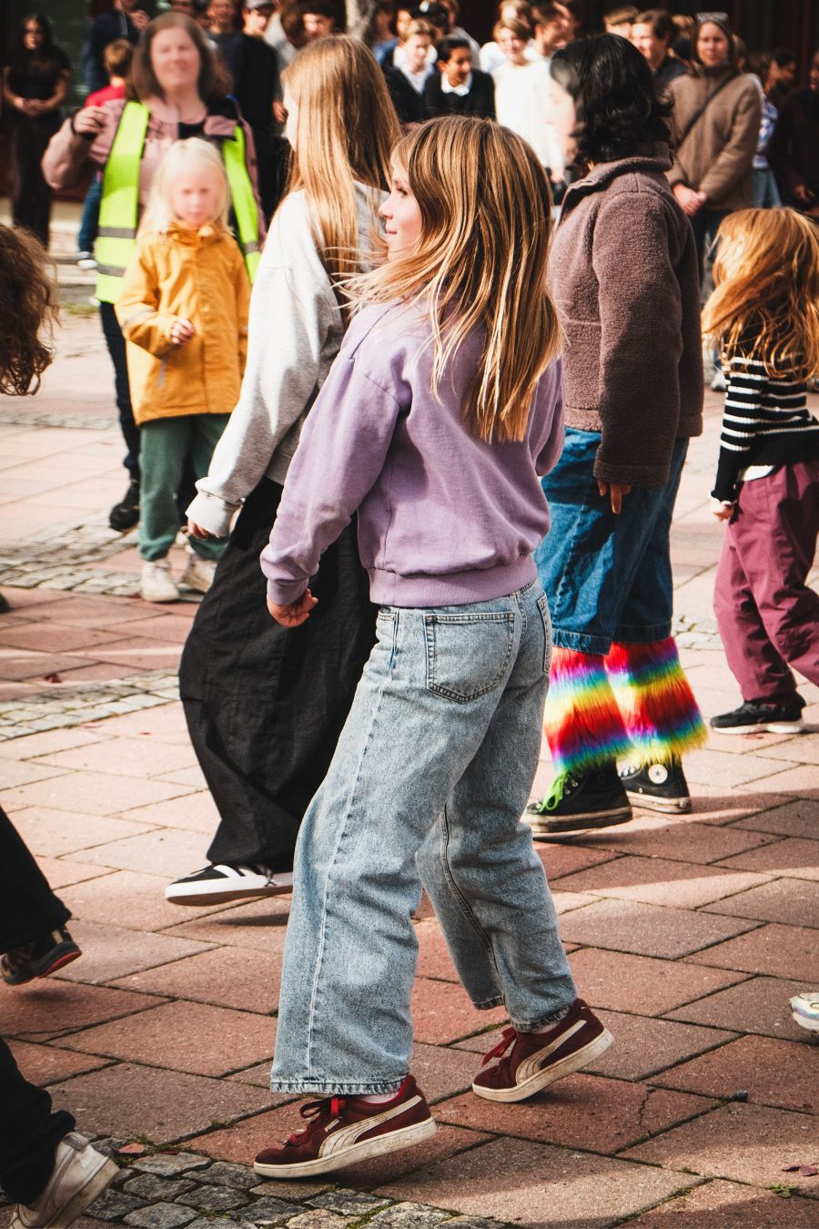 A large crowd of children and adults dance together outdoors on a paved square, wearing a variety of colourful jackets, jeans, and shoes, including a pair of rainbow-coloured fuzzy leg warmers. A girl in a lilac sweatshirt is seen mid-movement in the foreground.