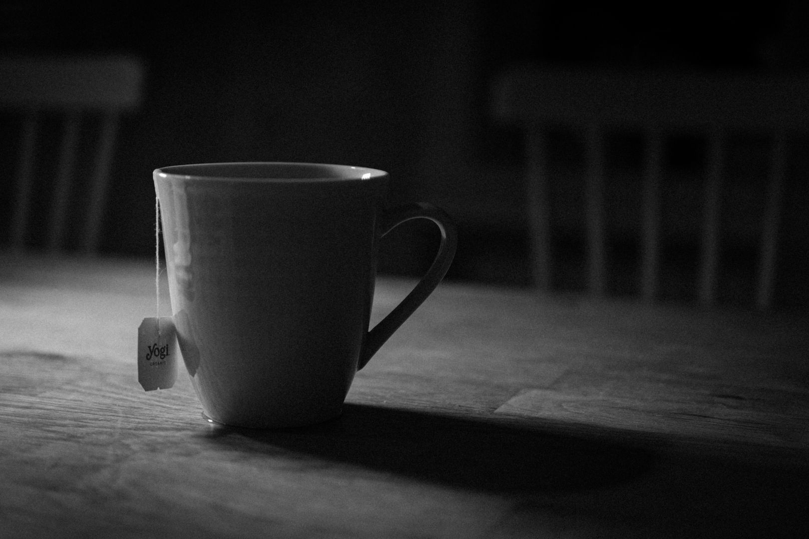 A black and white image of a mug with a tea bag tag hanging over the side, standing on a wooden table. Soft light falls across the mug and table, with two blurred chairs visible in the dim background.