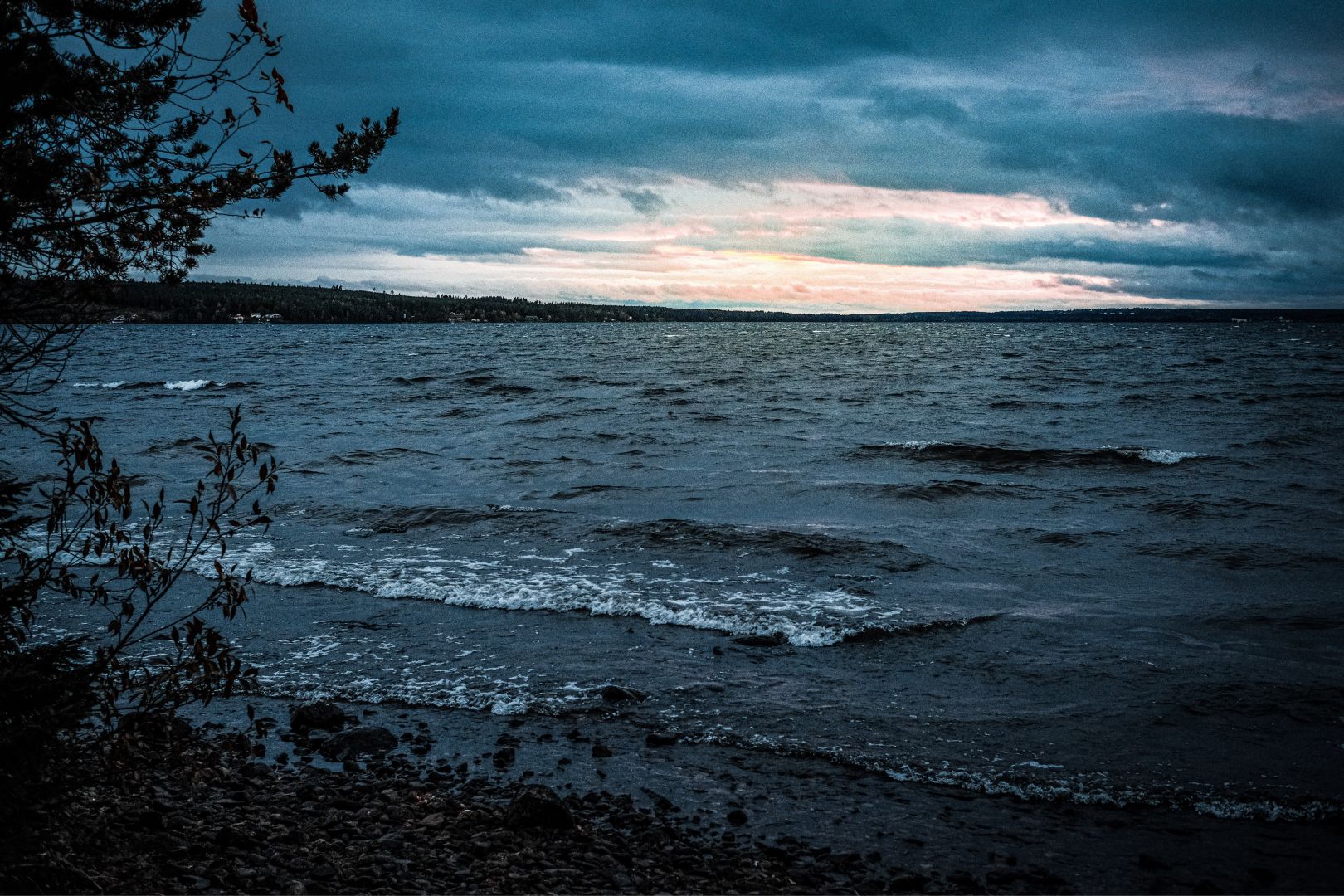 A dark, moody image of a lake at dusk, with waves washing onto a rocky shore in the foreground. Heavy clouds fill most of the sky, with a narrow band of warm light glowing at the horizon.