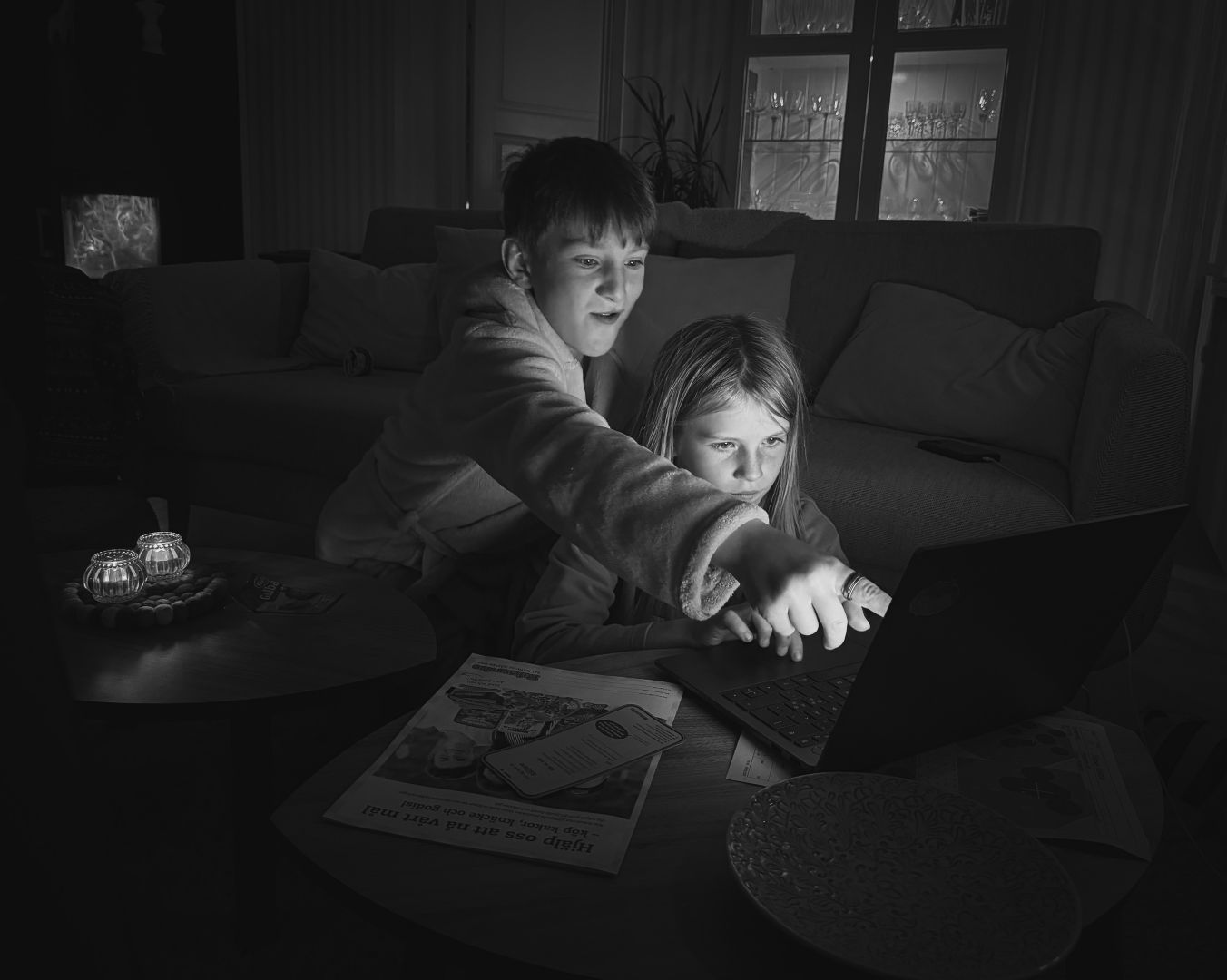A black and white image of two children looking intently at a laptop screen on a coffee table in a dimly lit living room. The older child points at the screen while the younger sits closer, focused on typing. Soft ambient light comes from a lamp and glass cabinet in the background.