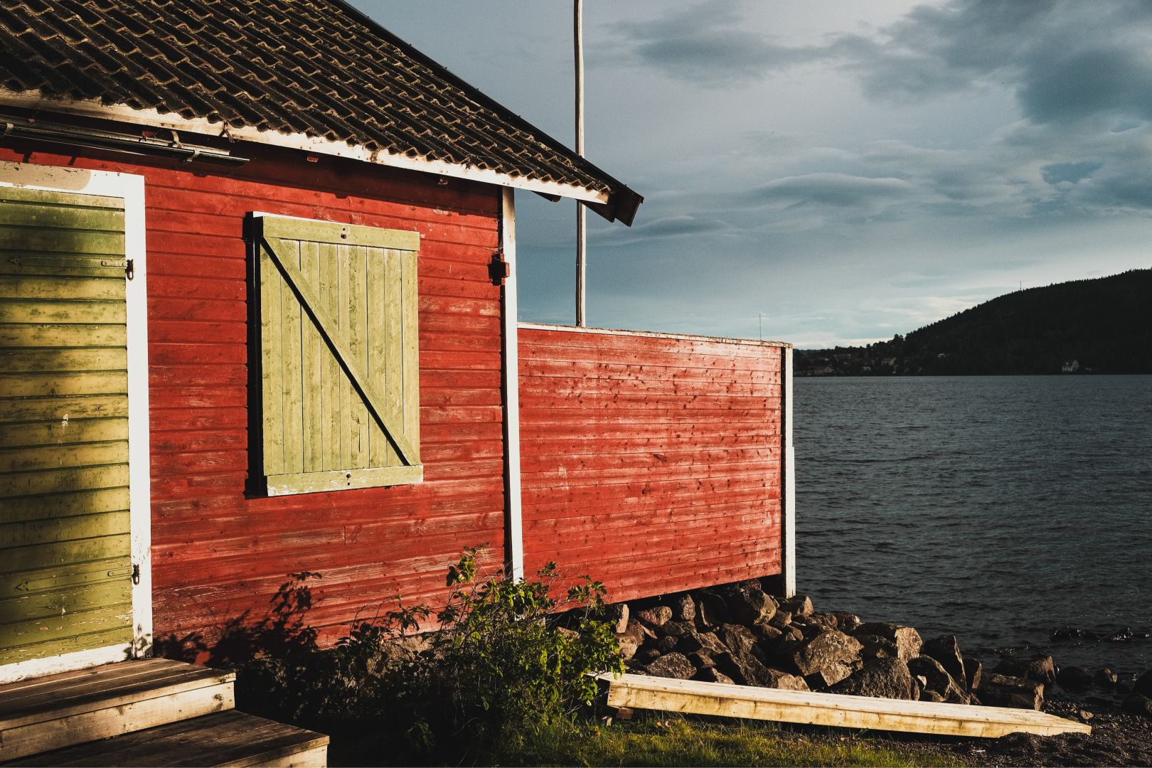A traditional red wooden boathouse with green shutters and a dark corrugated roof stands beside a rocky shoreline, overlooking a calm lake under a heavily clouded evening sky. Low forested hills line the far shore in the distance.