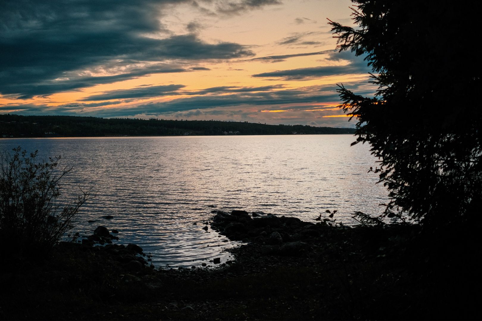A calm lake reflects the soft pastel colours of a sunset sky filled with layered clouds. Dark silhouettes of trees and bushes frame the rocky shoreline in the foreground, with a low forested landscape stretching along the far shore.