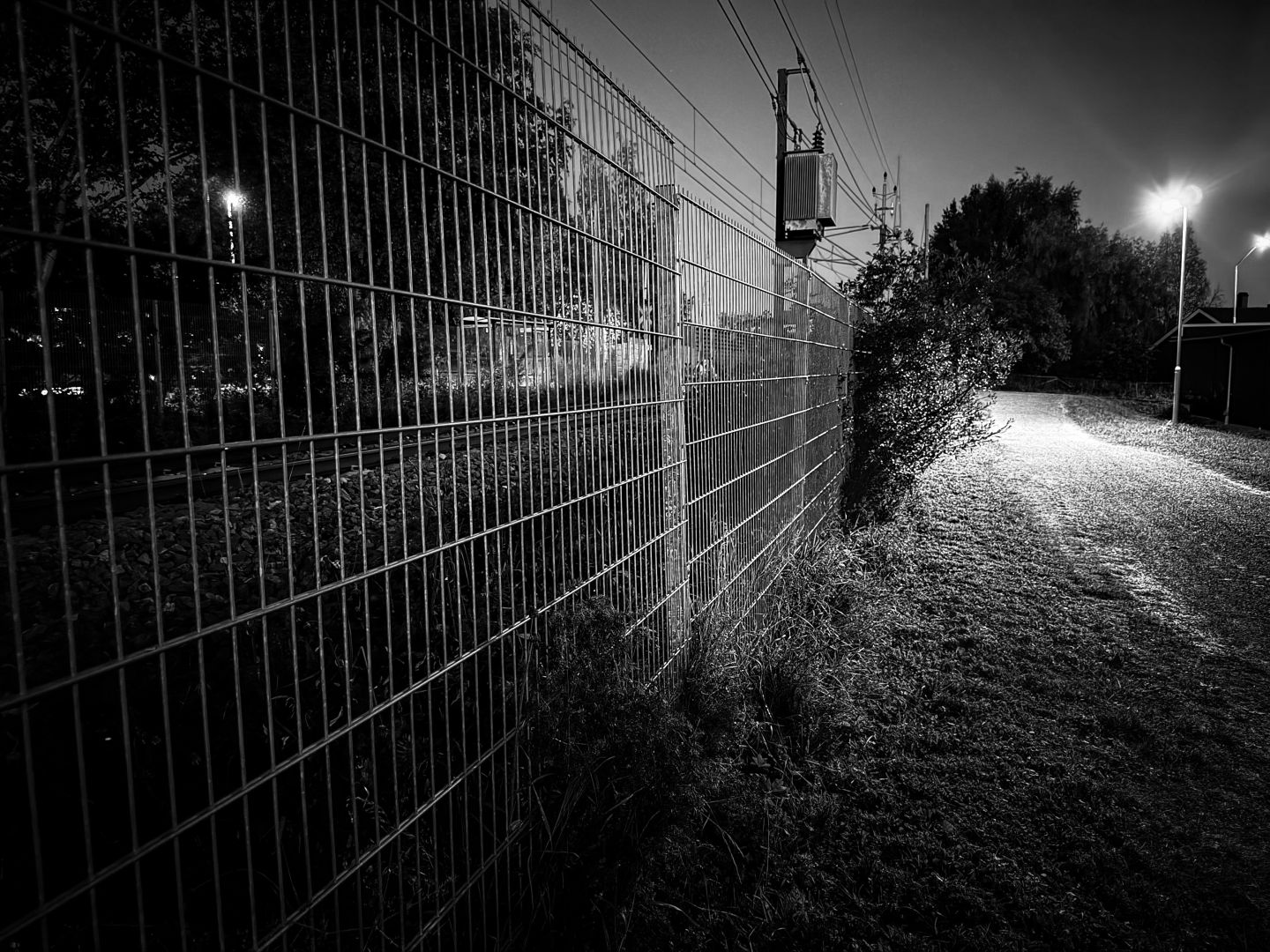 A black and white nighttime image of a tall wire fence running alongside a gravel path illuminated by a bright streetlight. Power lines and a transformer are visible above the fence, with dark trees and distant lights fading into the background.