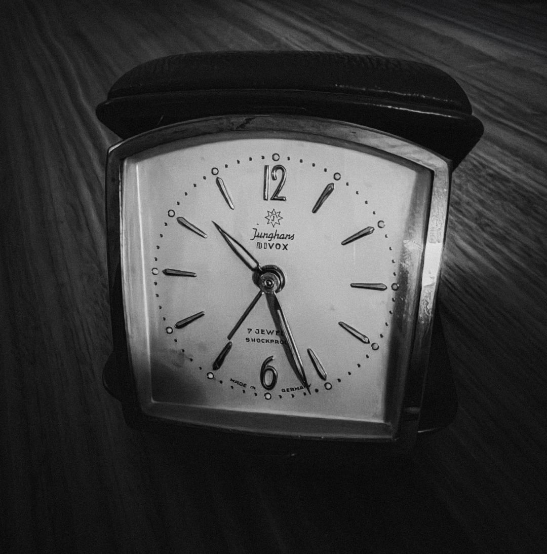 A black and white close-up of a vintage Junghans travel alarm clock in a leather case, showing its white face with black numerals and hands set to around 8:30, resting on a wooden surface.