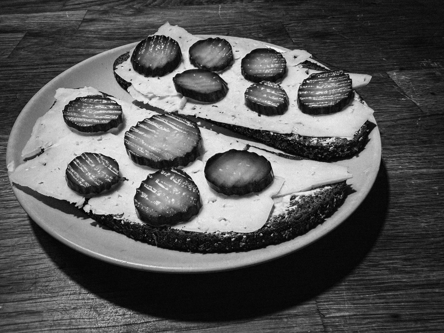 A black and white close-up of an open-faced sandwich on dark rye bread, topped with slices of cheese and pickled cucumber, arranged on a round plate. The photo is taken with an ultra-wide lens, giving a slightly curved perspective of the wooden table beneath.