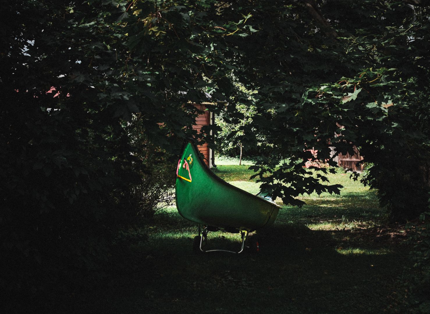 A green canoe rests on a stand in a shaded garden area, tucked beneath overhanging tree branches. Dappled sunlight filters through the leaves onto the grass, with a wooden shed and fence visible in the background.