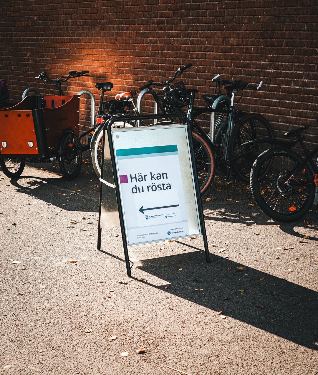 A sandwich board sign reading "Här kan du rösta" (Here you can vote) with an arrow, standing on a paved area in front of a brick wall lined with parked bicycles, including a cargo bike. Warm afternoon light casts long shadows across the ground